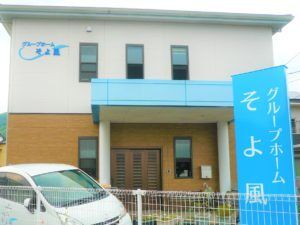 2階建ての建物の1階部分は木目調、2階部分は白で、外壁にはホーム名が書かれている。玄関には屋根が付き、そばには車が止められている。