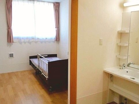 介護ベッドが置かれたフローリングの部屋にはレースカーテンとカーテンが付いた窓があり、洗面所も付いている。
