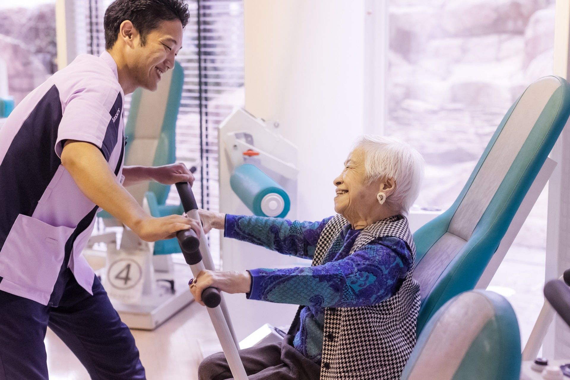 赤い椅子が平行棒の前に並べられ、高齢者たちが並んで立ち、スタッフの指導を受けながら足の上げ下げをしている。