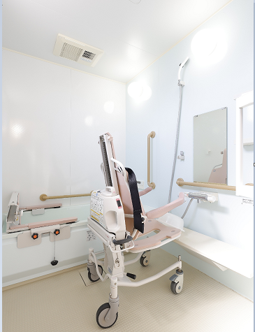 車いすの高齢女性と付き添いの高齢男性が花が咲く明るい道を笑顔で散歩している。そばにはエプロンを付けた若い女性スタッフもいる。