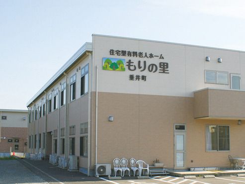 広い駐車場があり、日当たりのいい場所に建っている2階建ての建物はエル字型。1階部分がベージュ、2階部分が白い外壁。