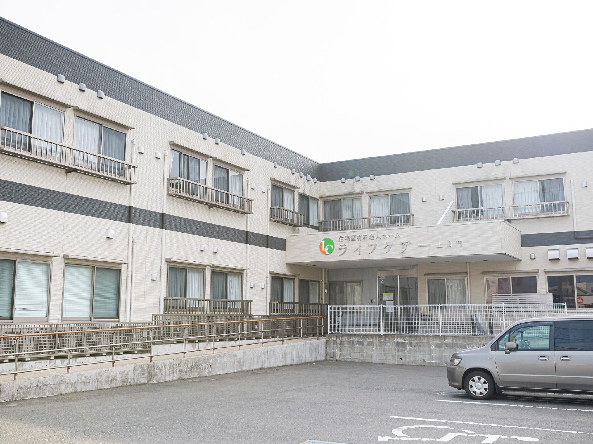 手すり付きのスロープが付いた玄関には屋根も付いている。駐車場が付いた2階建ての建物はエル字型。