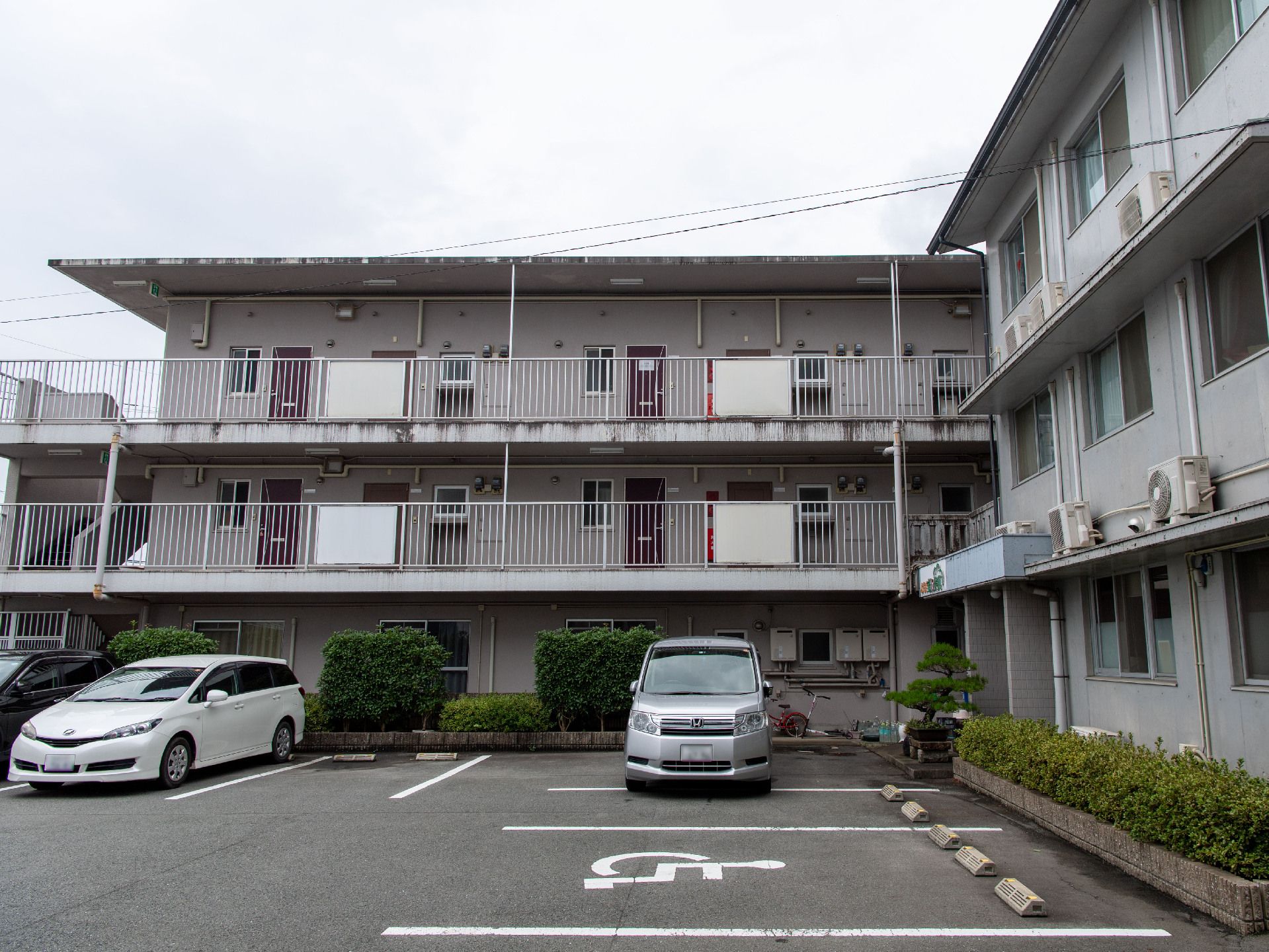 L字の灰色の壁の建物の前にある車が止まった駐車場