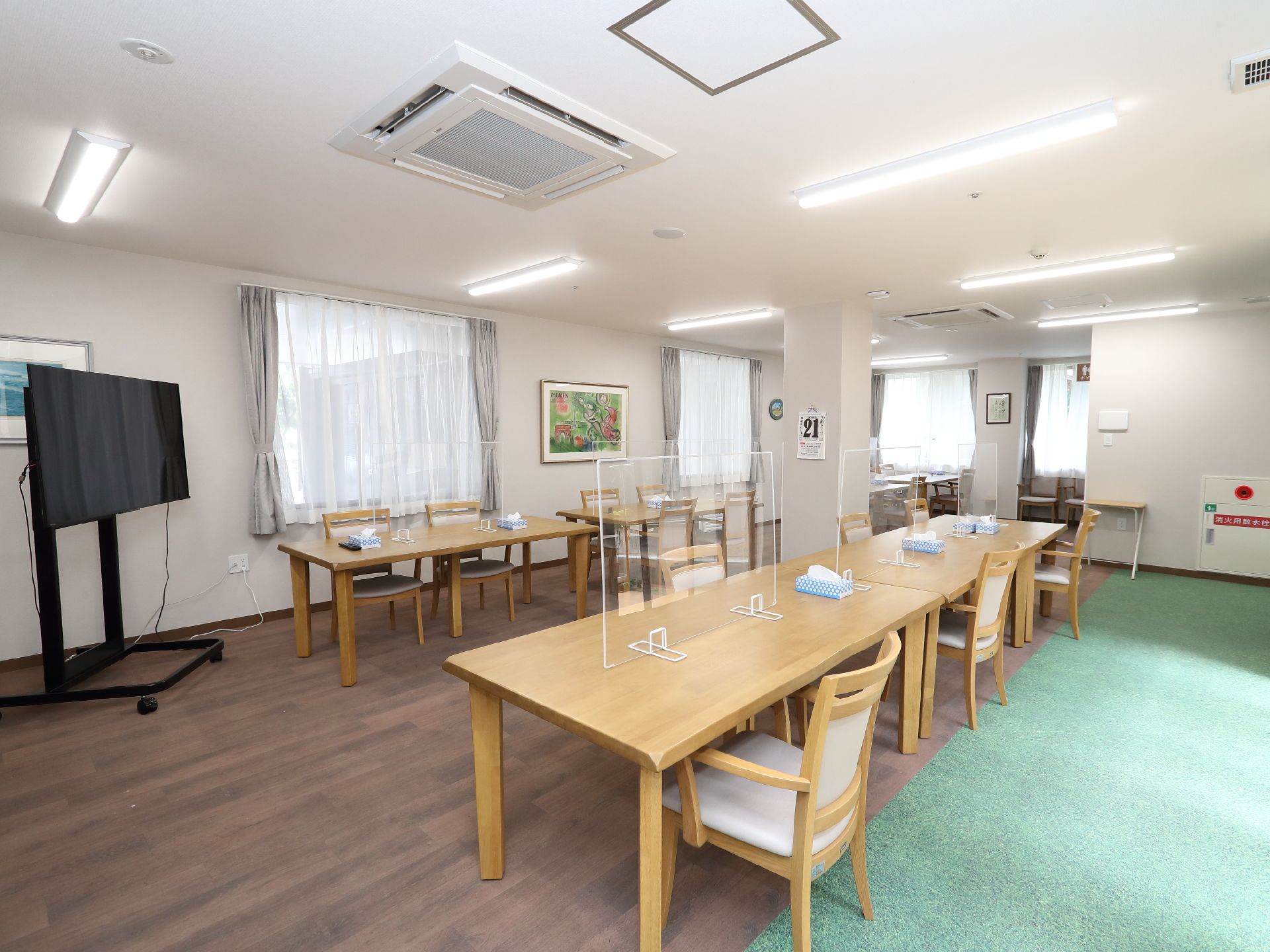 透明な仕切りが付いた木製の机と椅子が並べられた窓ガラスが複数配置された広間