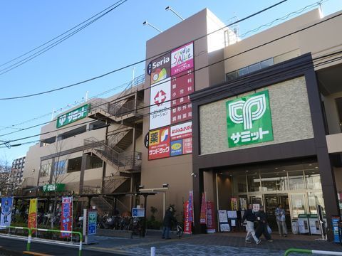 スーパーの建物がある。壁面には、ドラッグストアやクリニックなどの案内看板が貼られており、店舗前にはさまざまなのぼりが立てられている。