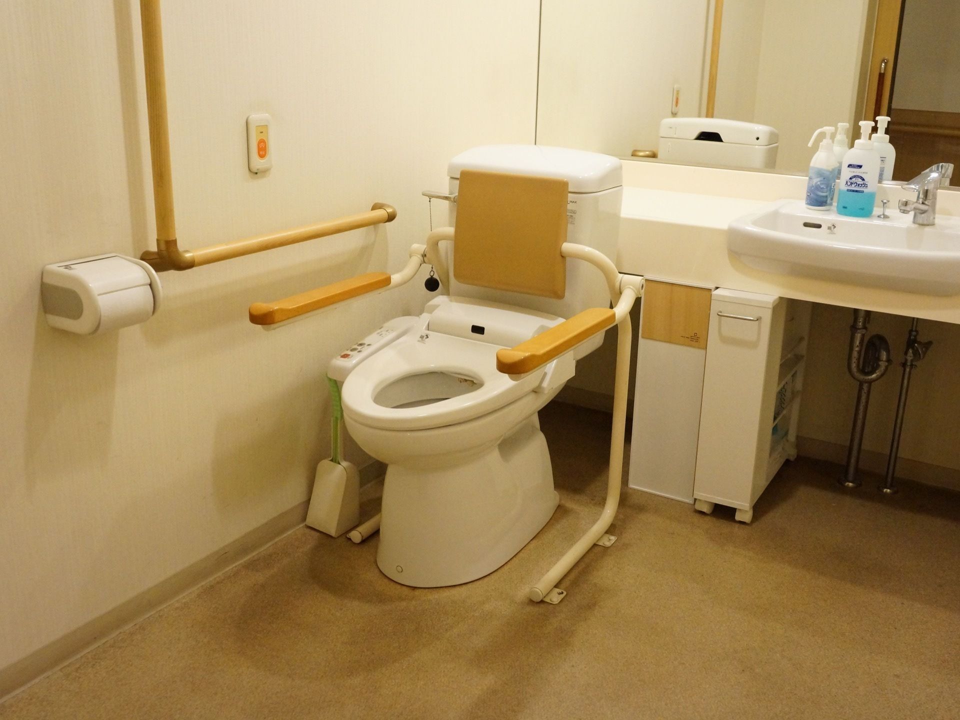 広い個室トイレである。洗面台下にはキャスター付きの小さな収納棚が置かれており、鏡前にはハンドソープや消毒液が置かれている。