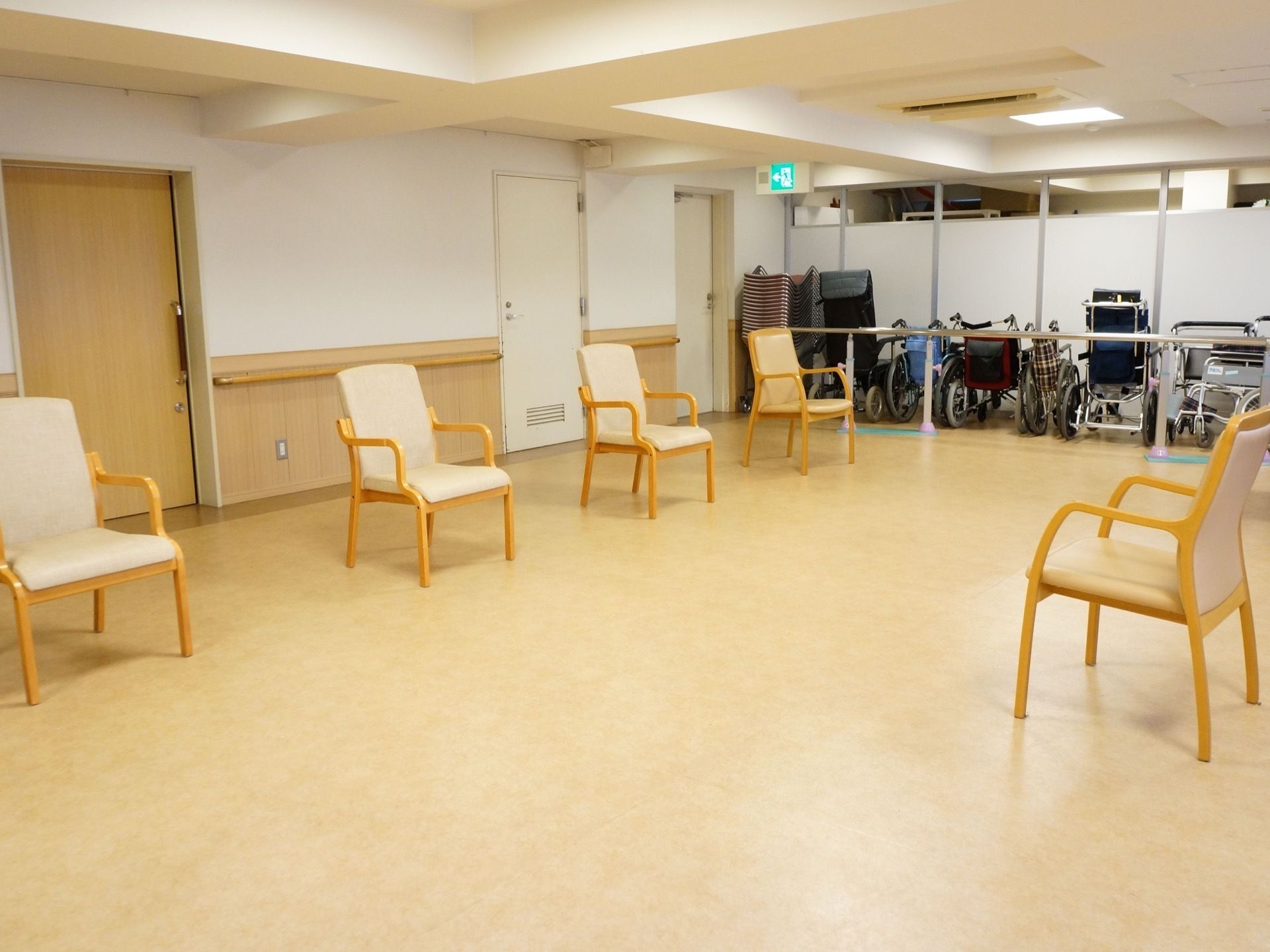 奥にさまざまのタイプの車椅子が複数置かれている。壁には木目調のスライド式の扉と非常口があり、手すりが取り付けられている。