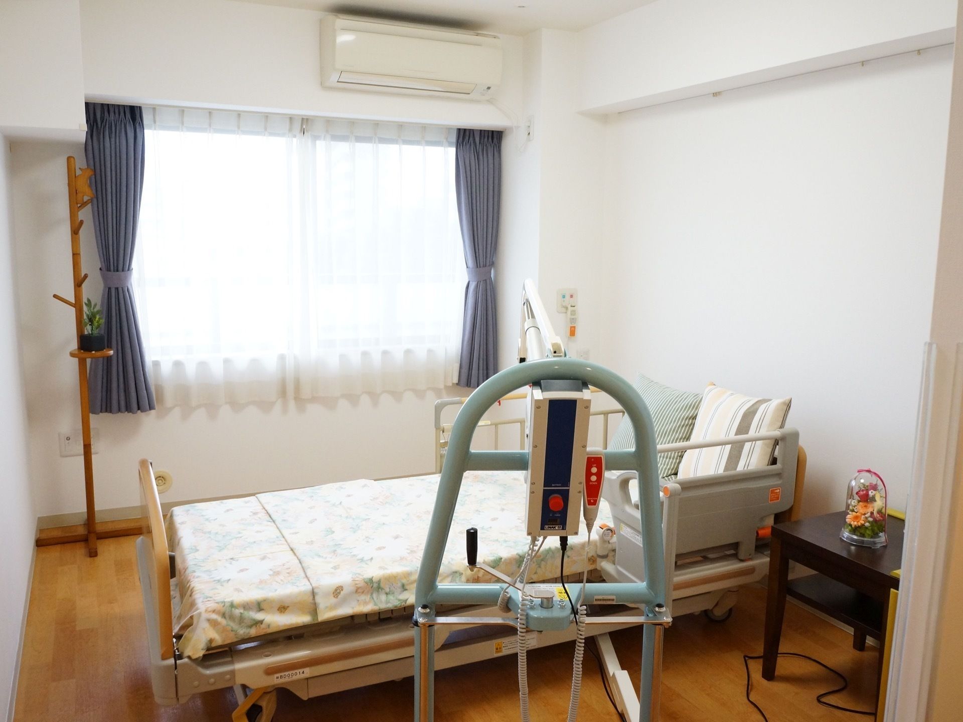 日当たりの良い居室で、窓にはレースと青色のカーテンが取り付けられている。壁にはエアコンが設置されている。