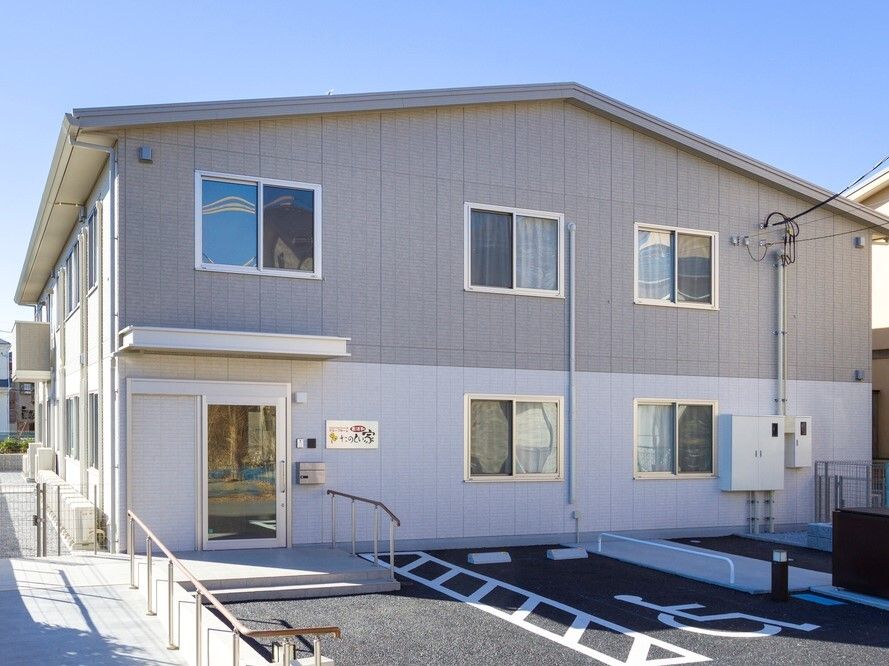 2階建ての建物の1階部分は白、2階部分はグレーで、駐車場もあり、玄関には手すり付きのスロープが付いている。