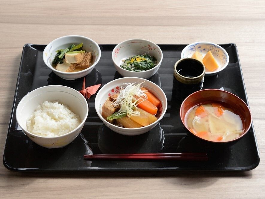 白いご飯に味噌汁、煮物や野菜の和え物などがそれぞれ器に盛られて黒いトレーに並べられている。