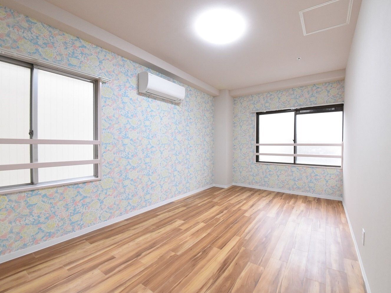 パステルカラーの花柄のような壁紙が貼られたフローリングの部屋。カーテンの付いていない窓が二つ設置されている。