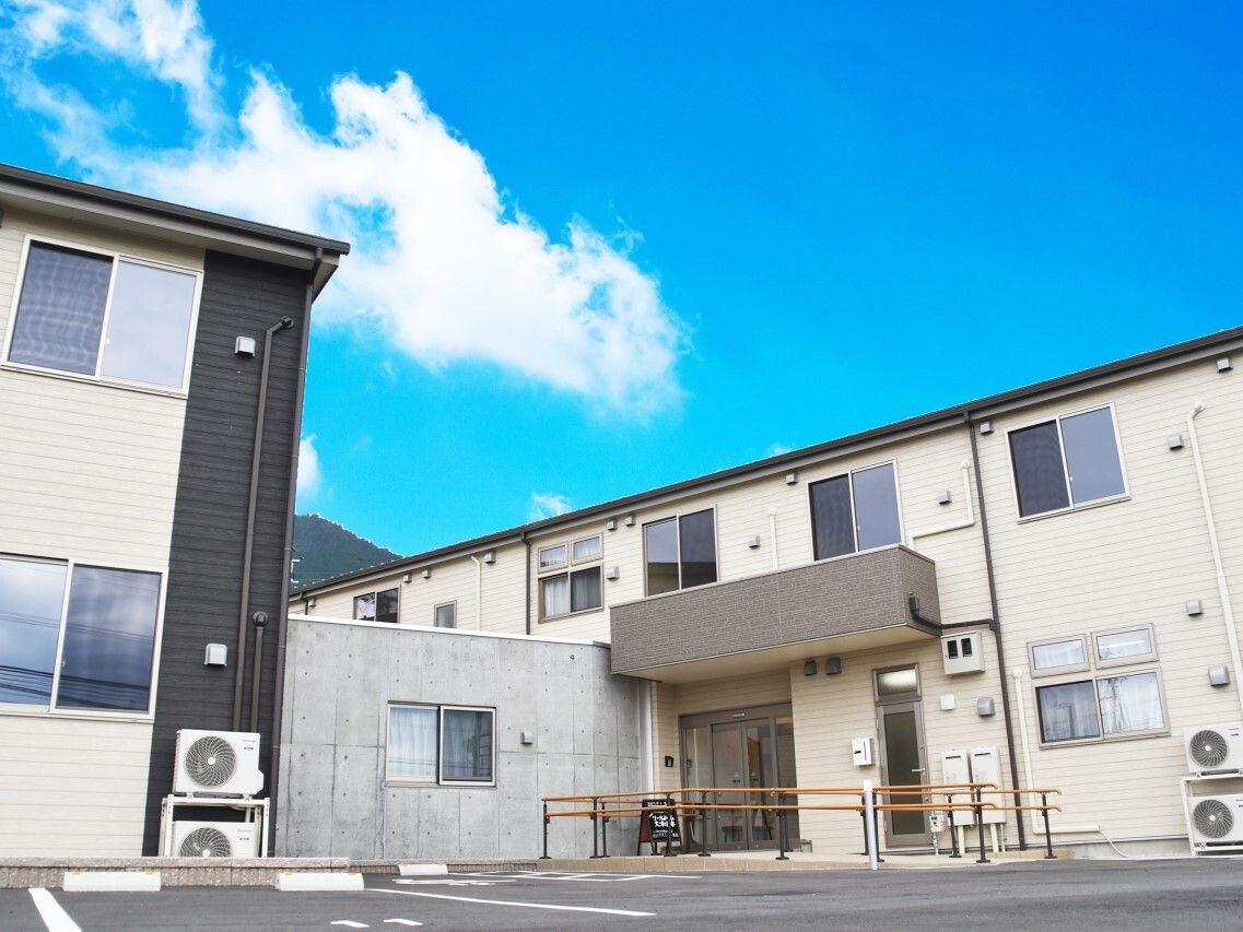 手すりが付いた玄関には屋根もあり、自動ドアが設置されている。2階建ての建物はベージュ系の外壁で横に長いデザイン。