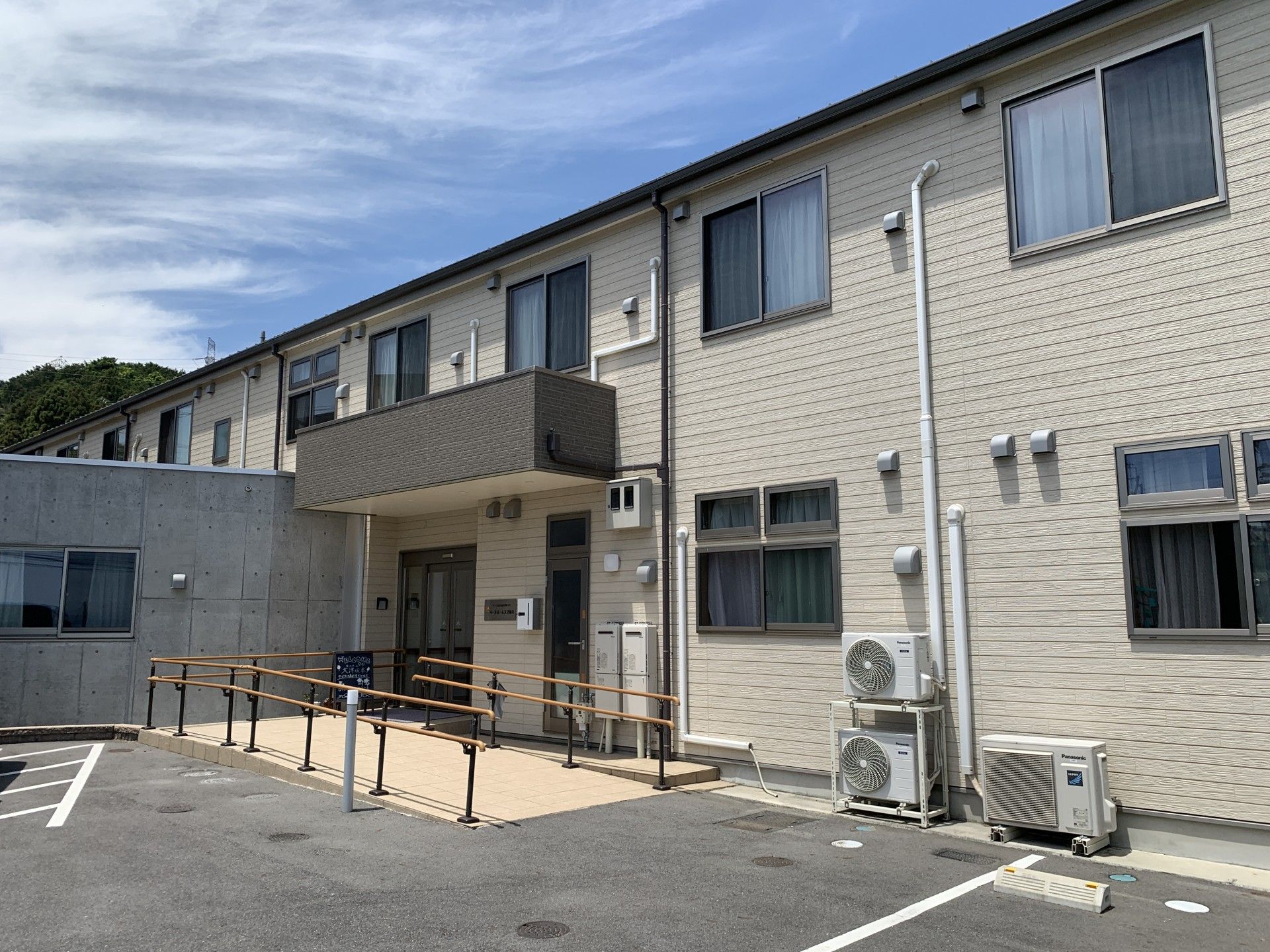 青い空の下に建っている2階建ての建物。玄関には手すりが付き、敷地内には駐車場があり、広々としている。