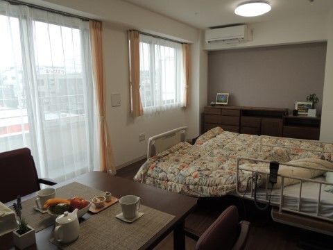 エアコンが完備された明るく広々とした居室である。リモコンとフェンスが付いた介護ベッドが２台置かれている。