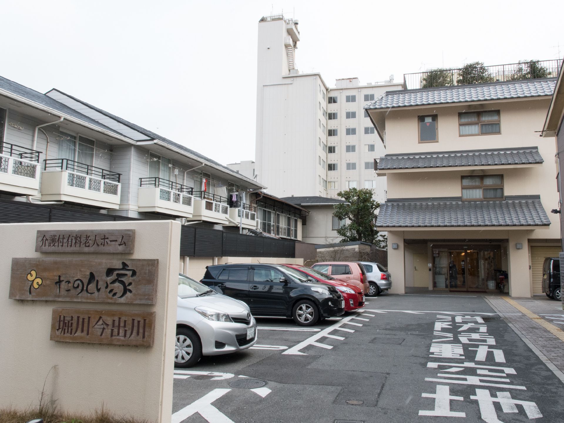 左手前に施設名の看板がある。駐車場には自動車が数台駐車してある。地面に白く「消防活動空地」の文字がある。