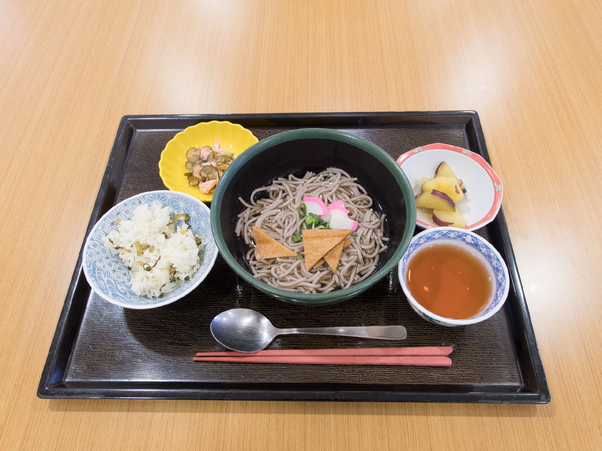 麺類を中心とした食事が黒い盆に載っている。小鉢が4つあり、1つは麺の付け汁、3つは副菜である。スプーンと箸が添えられている。