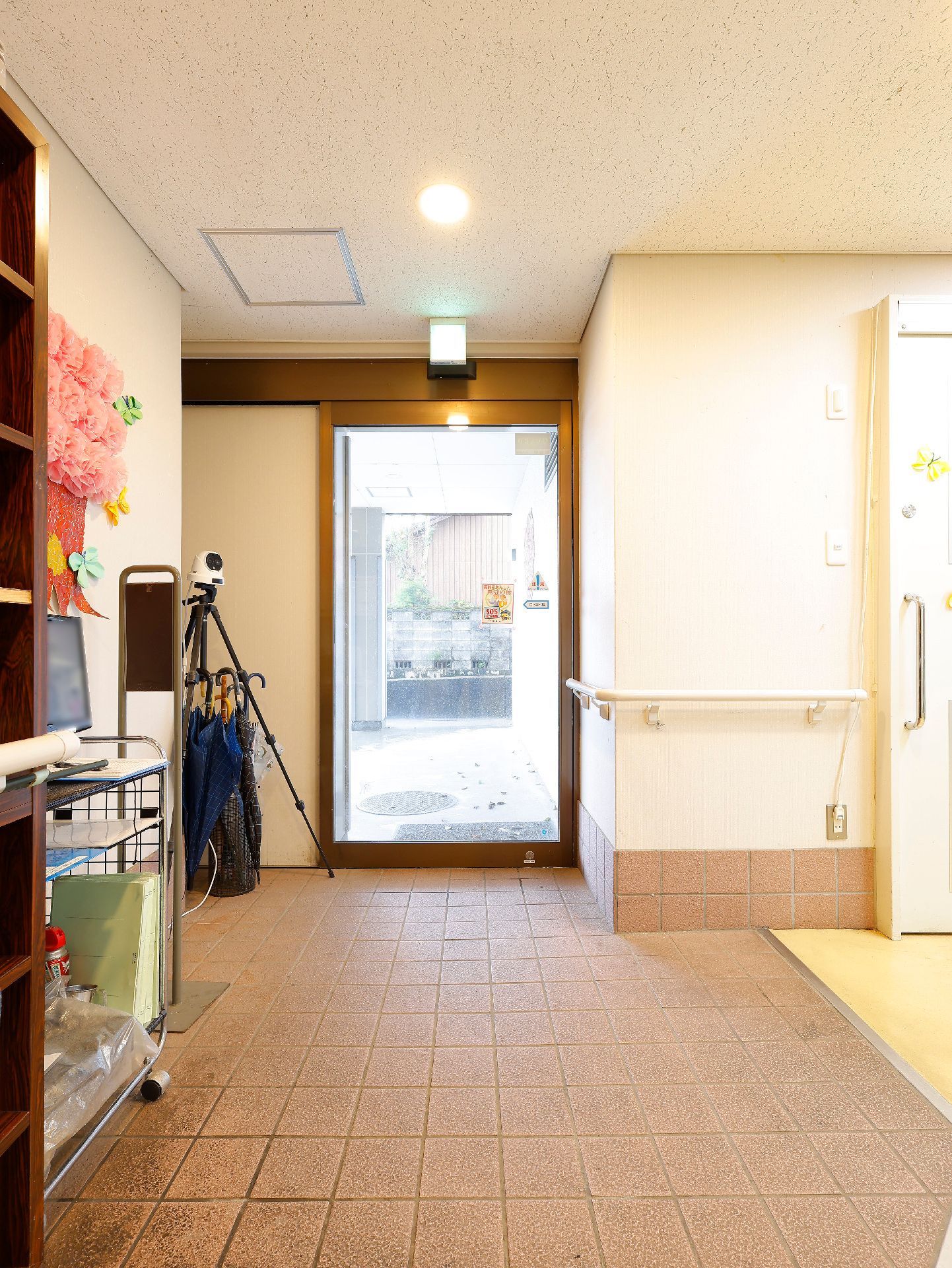 ピンクのエプロンを付けた女性スタッフが黄色いカーディガンを着た高齢女性に寄り添っている。女性は車いすに座っている。