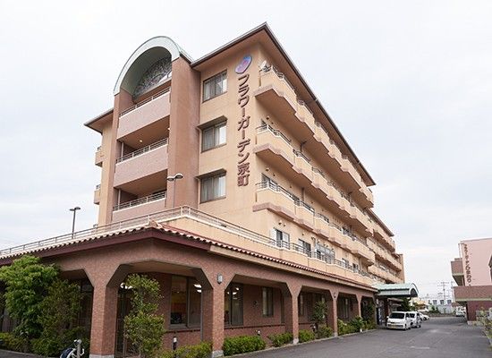 ピンクのエプロンを付けた女性スタッフが高齢女性が薬を飲みこむのを待っている様子。手には水の入ったコップがある。