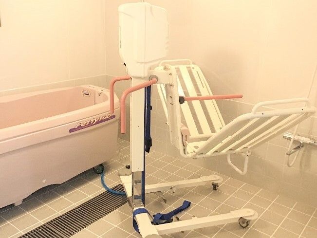 ストレッチャータイプのリフトがピンクの浴槽型の機械の隣に置かれている。タイル張りの浴室。