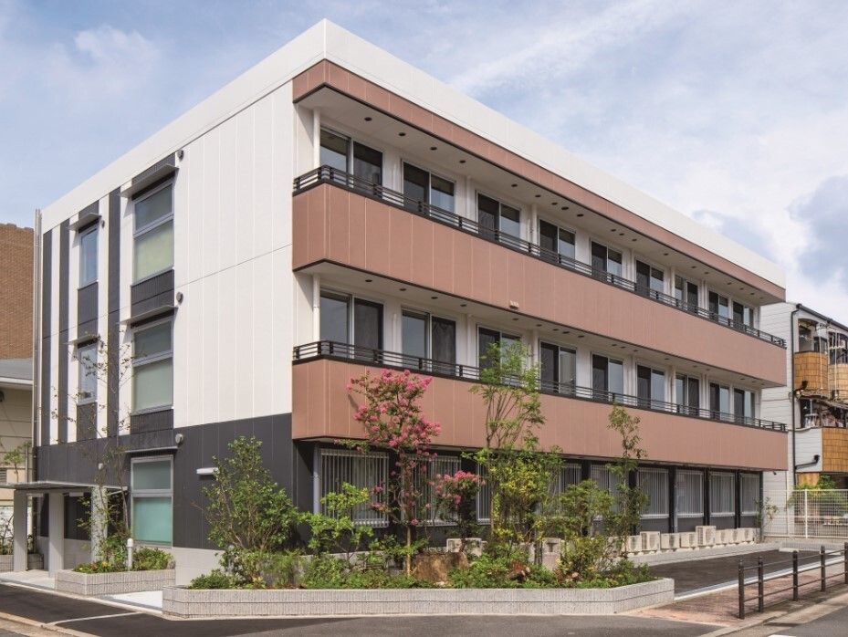 各フロアにブラウンのベランダが付いているモノトーンの3階建ての建物。周りにはビルようなものがある。