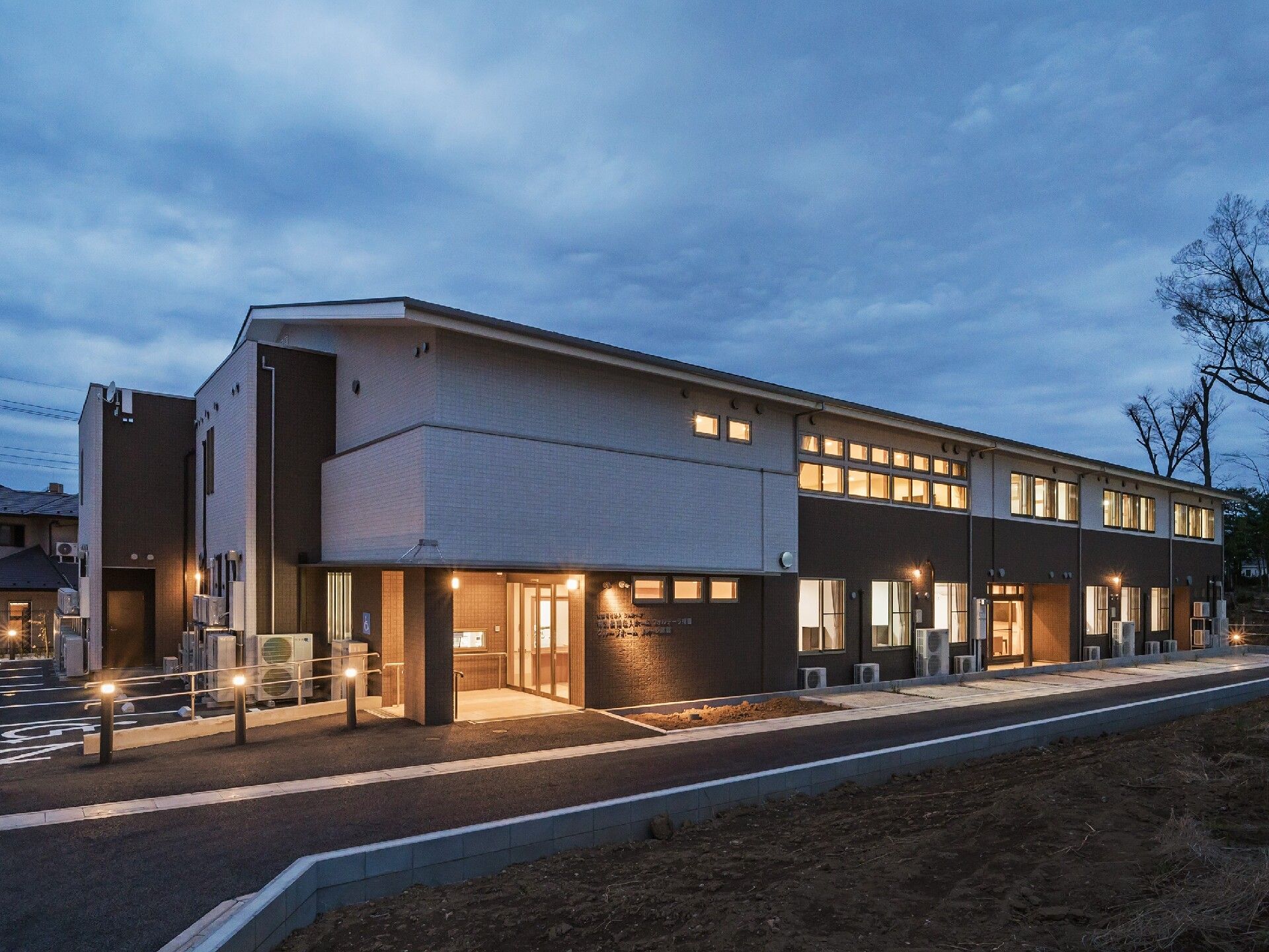 夕暮れ時になり、館内に明かりが灯った横に長い建物。玄関先には外灯が点き、手すりが設置されている。