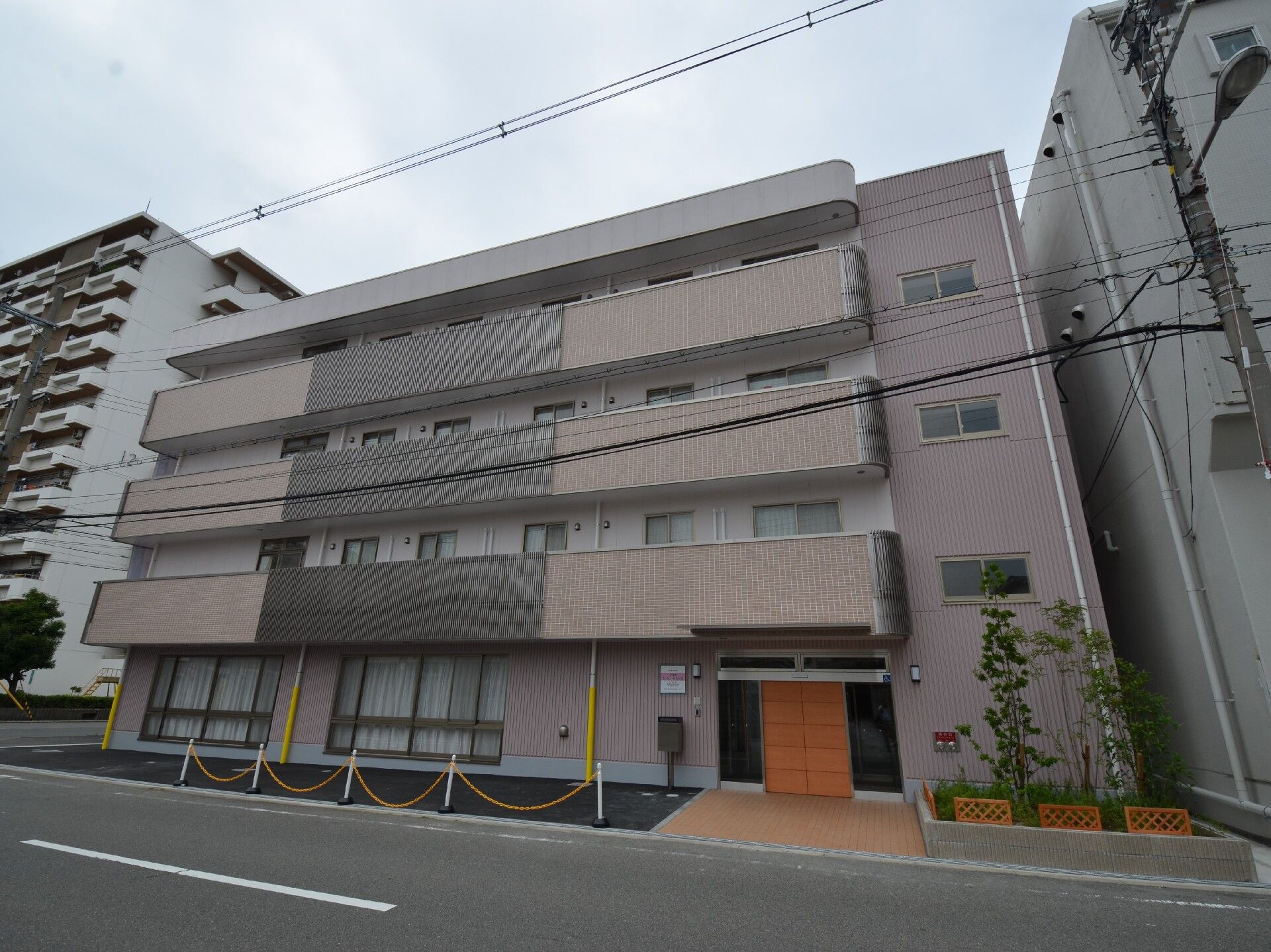4階建ての建物はブラウン系で各フロアにベランダがあり、道路に面している。周りにもいくつか建物がある。