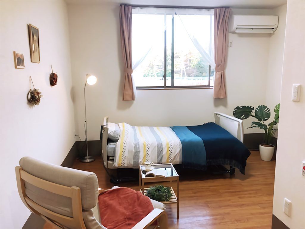 寝具付きのベッドが置かれたフローリングの部屋には一人用の椅子や観葉植物が置かれている。