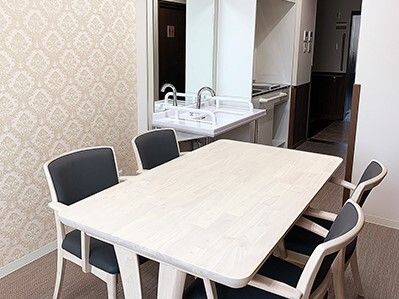白いテーブルには黒い椅子が4脚セットされ、奥には洗面所とキッチンがあり、フロアはフローリング。
