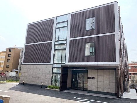 駐車場が付いた3階建ての建物の1階部分はベージュ、2階よりも上はダークブラウンで、玄関のそばには名前が書かれている。