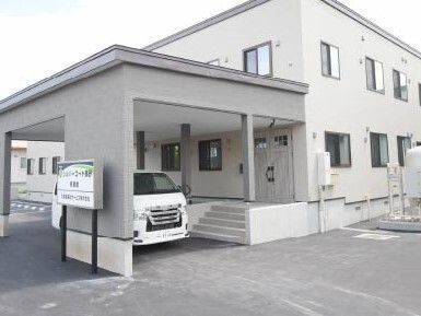 ライトグレーの外壁の2階建ての建物。玄関には手すり付きのスロープと階段があり、車が止められている。