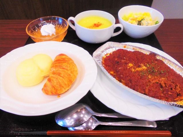 アルミ皿に入ったグラタンのような料理とパンが2種類、スープ、ゼリー、野菜の料理などがそれぞれ盛られている。
