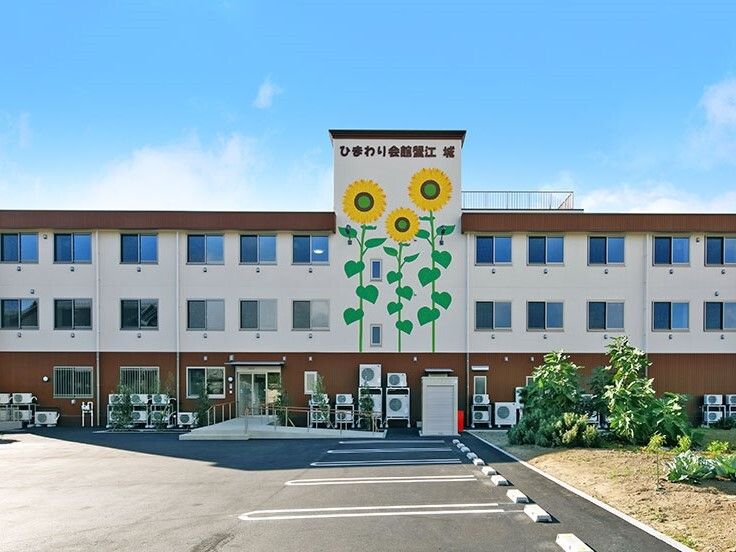 白とブラウンの３階建ての建物。ヒマワリの絵が描かれた外壁が印象的。駐車スペースも広い。