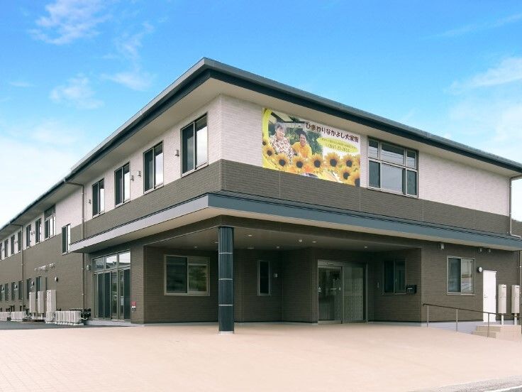 2階建ての建物は1階部分がダークグレー、2階部分が白で、玄関には屋根が付き、手すり付きのスロープもある。