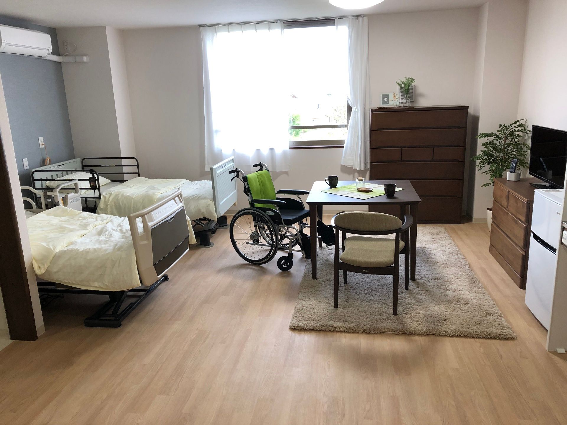 寝具がセットされたベッドが2台置かれたフローリングの広い部屋にはテーブルセットやテレビ、タンス、車いすなどがある。
