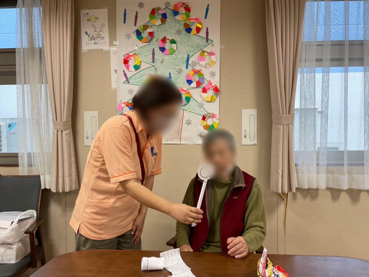 〇が描かれた棒をポロシャツを着た女性スタッフが高齢男性に手渡している。男性は座っている。