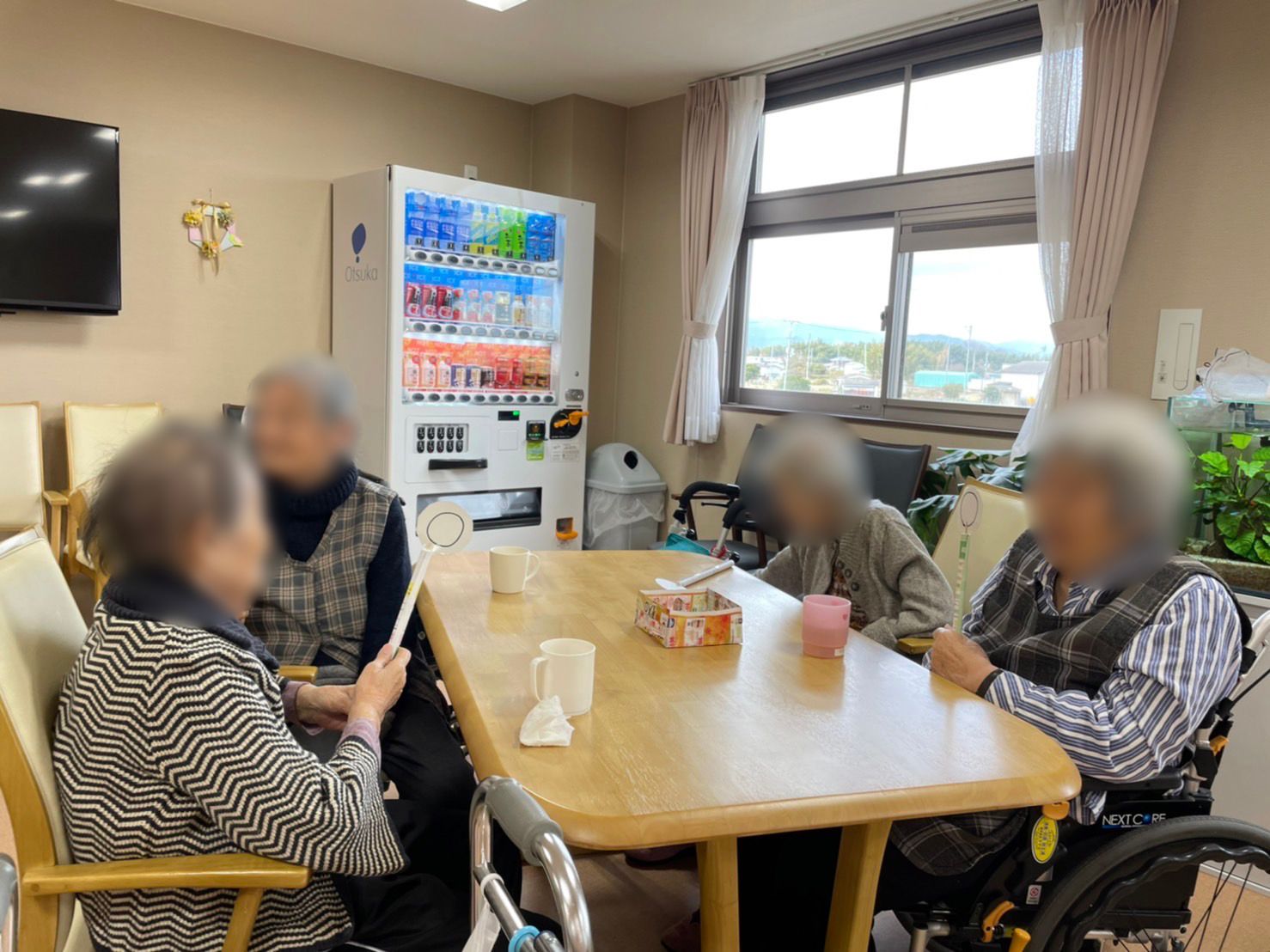 高齢女性４名がテーブルを囲んでいて、一人が〇と描かれた紙が付いた棒を持っている。奥には自動販売機もある。