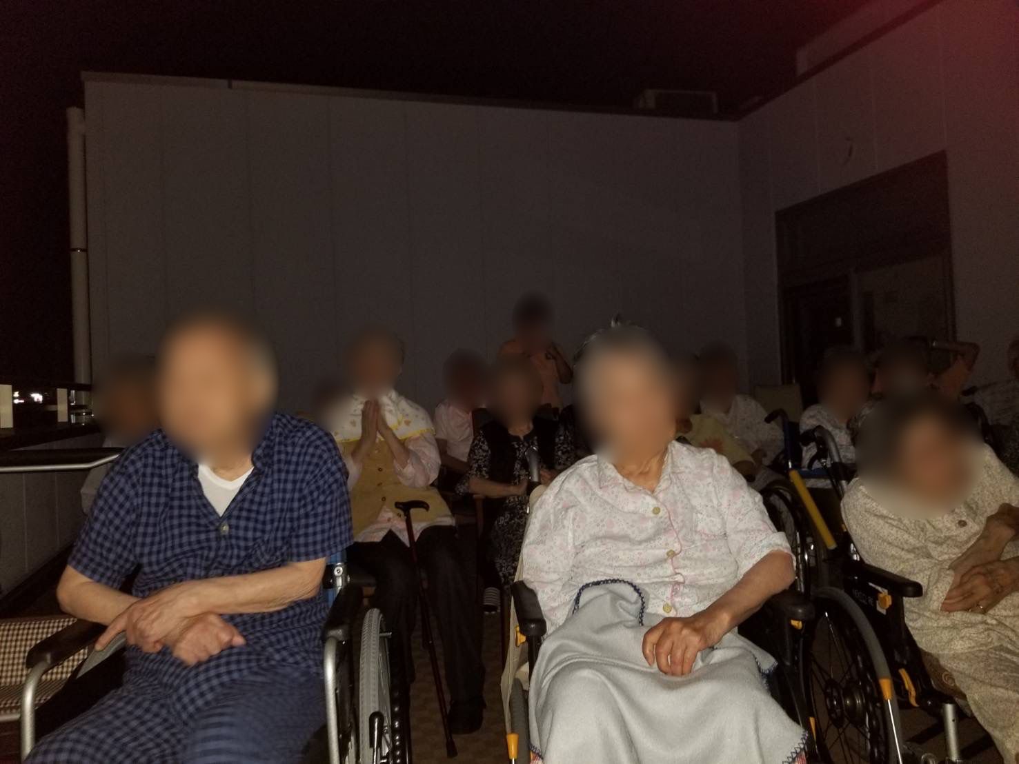 車いすに座った男女の高齢者たちが暗闇の中で座っている。麻痺がある人も数名いる。