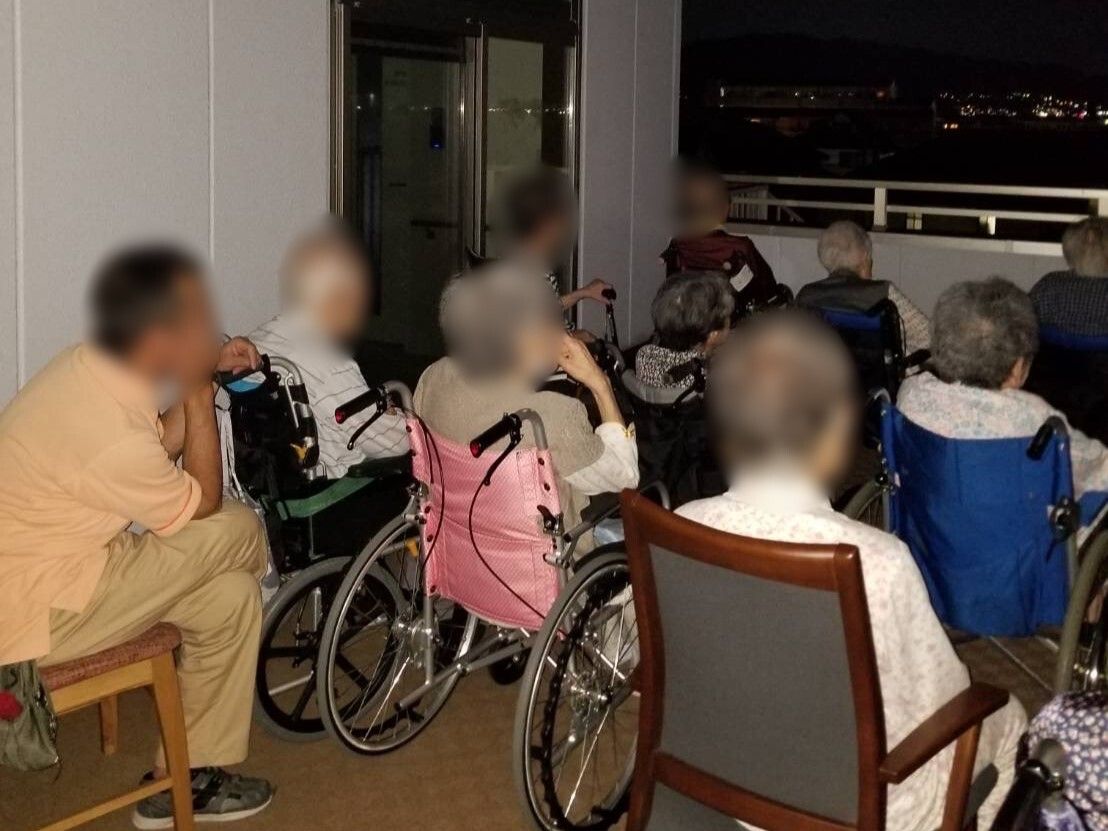 カーディガンを着た車いすの高齢女性が笑顔で目の前にいる女性が差し出す手を見ている。