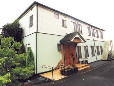 ベージュ系の外壁の２階建ての建物。敷地内には緑がたくさん植えられ、フェンスにはホームの看板が掛けられている。