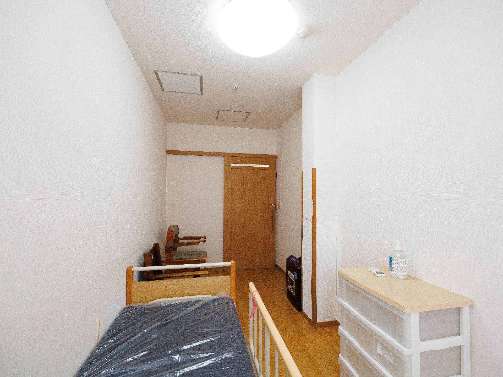 明るい雰囲気の道を高齢男性とピンクのエプロンを付けた女性スタッフに付き添ってもらいながら散歩している車いすの高齢女性。
