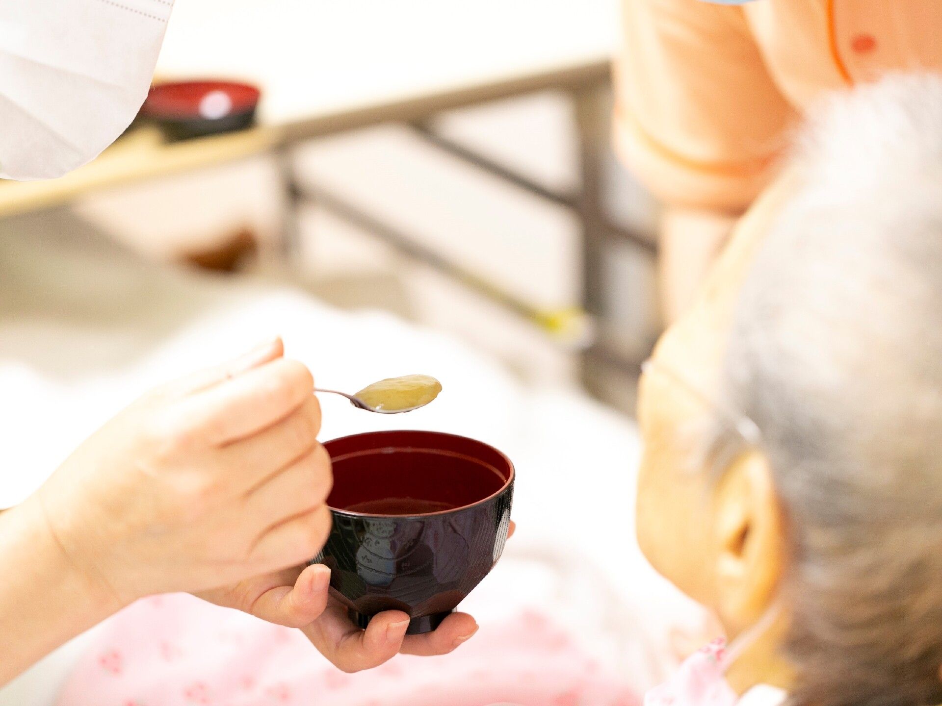 チューブを付けた高齢男性に銀の小さなスプーンで緑色のミキサー食を食べさせているスタッフの手。