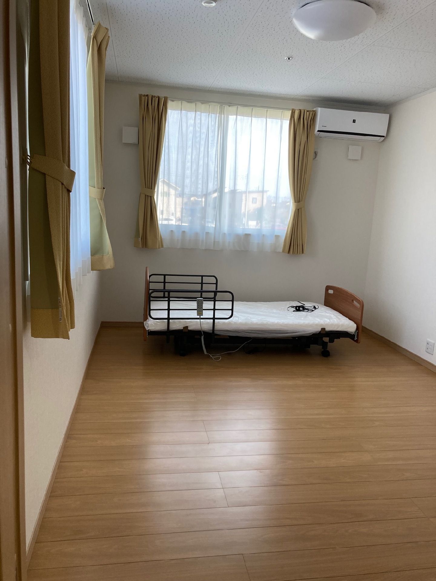 介護ベッドが置かれたフローリングの部屋にはエアコンが設置され、カーテンが付いた窓が２箇所にある。