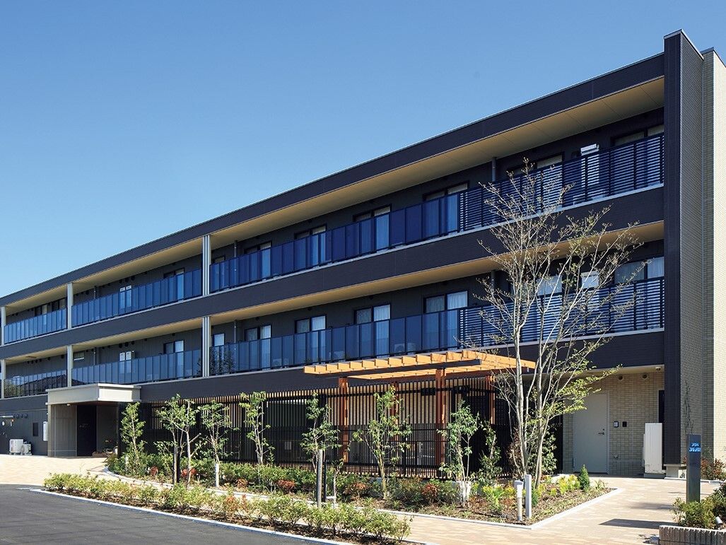 各フロアにベランダが付いたグレー系の建物は3階建てで、建物の周辺には草木がたくさん植えられている。