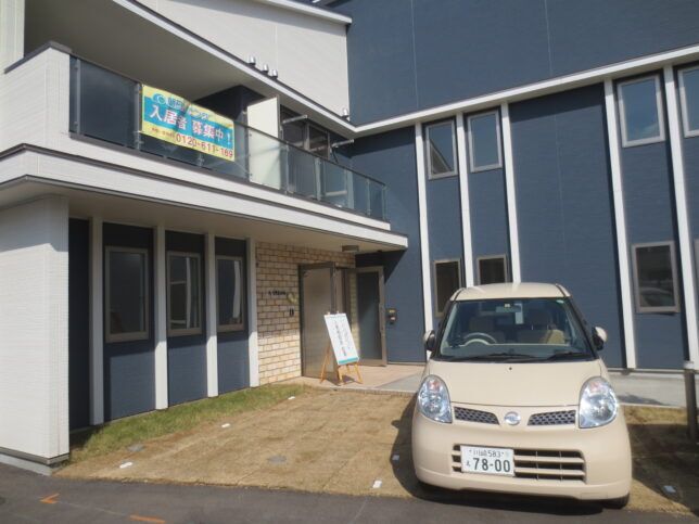 ベランダ付きの2階建ての建物の外壁はグレーと白で、建物の前には駐車場があり、車が止められている。