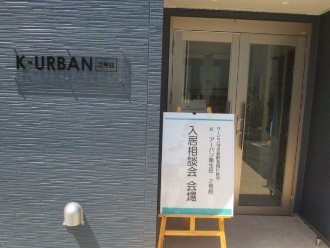 玄関の前には相談会と書かれた看板が建てられ、グレーの外壁には黒い文字で住宅名が書かれたプレートが貼られている。