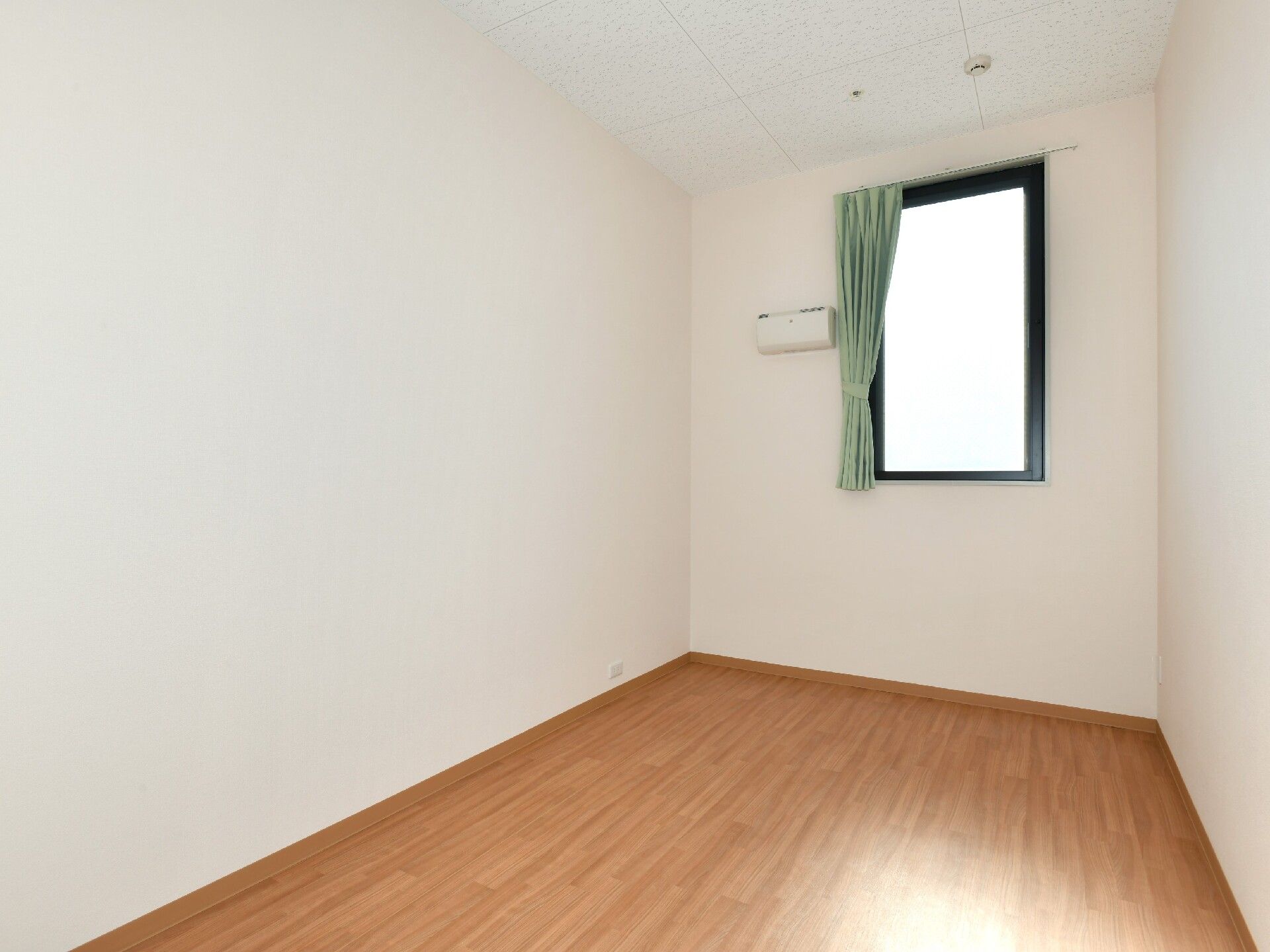 グリーンのカーテンが付いた小さな間があるフローリングの部屋には家具や家電はなく、エアコンもついている。