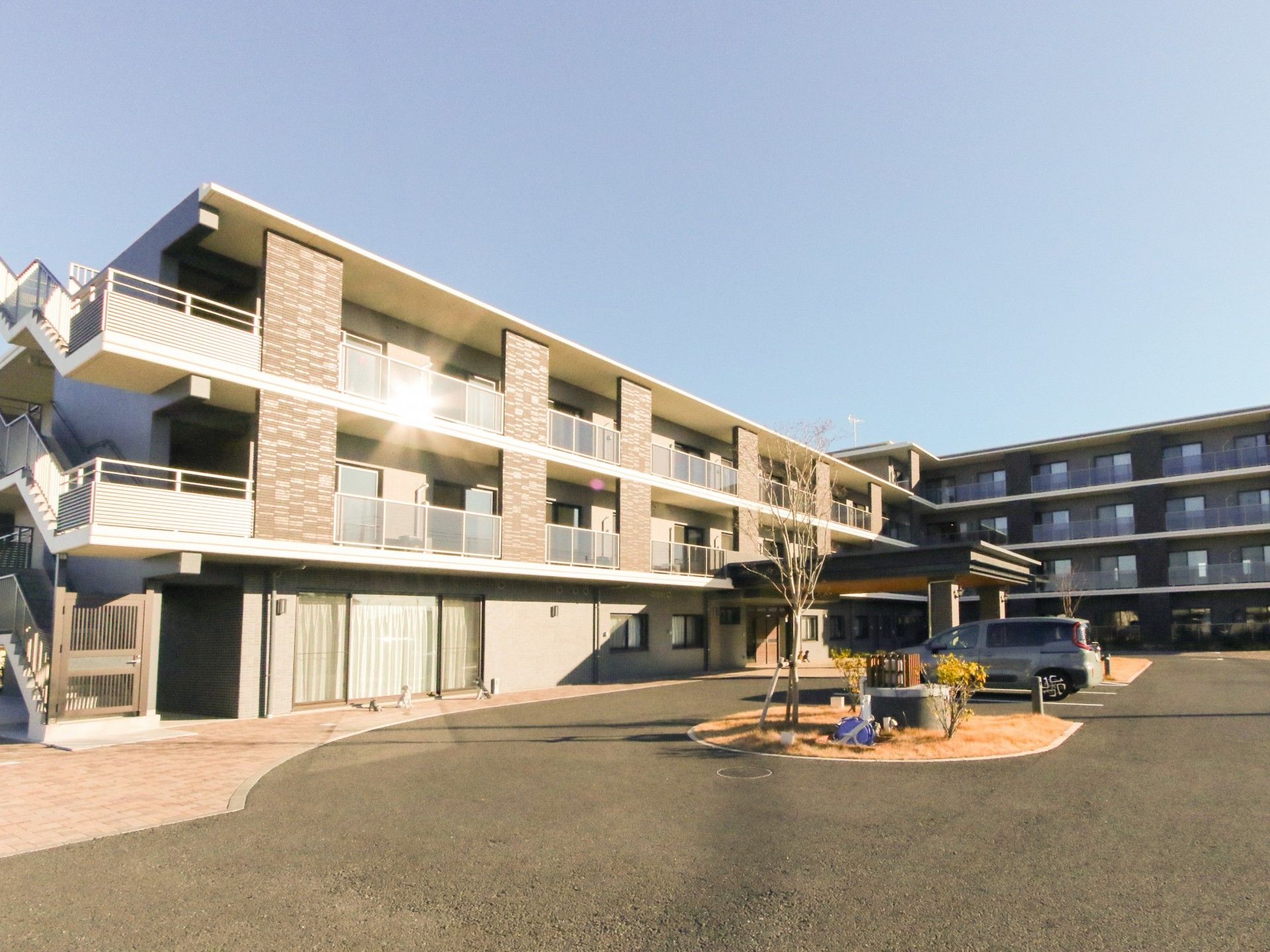 広い駐車場が付いた3階建ての建物はブラウン系で各フロアにベランダがあり、日当たりのいい場所に建っている。