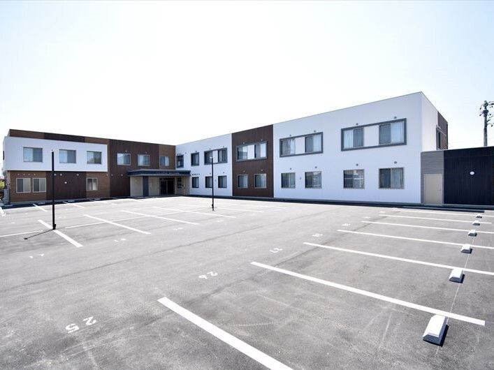 白線が引かれた広い駐車場が付いた２階建ての建物はエル字型で白とダークブラウン。玄関には屋根がある。