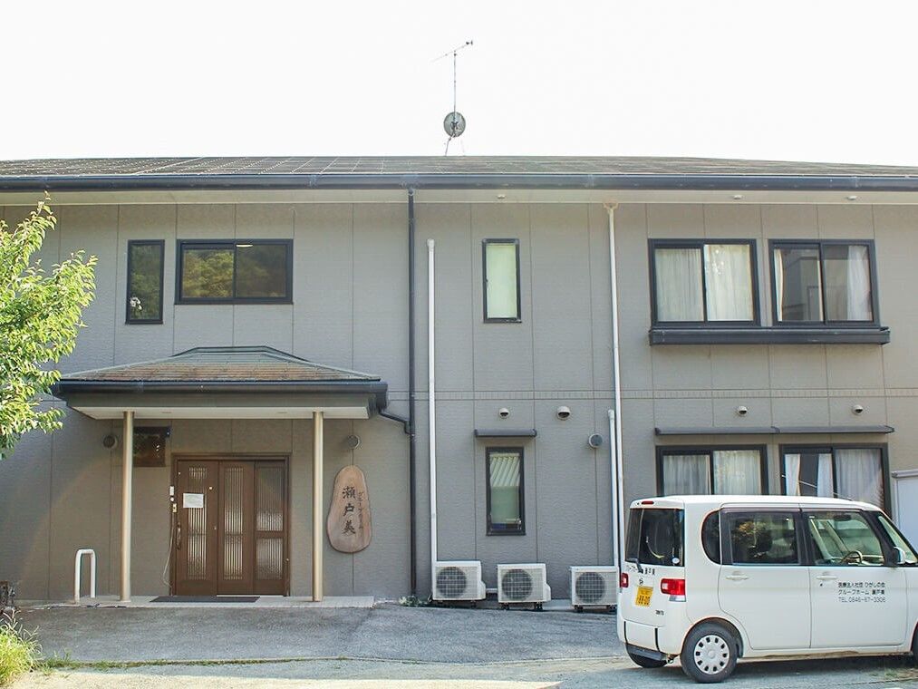 車が止められた駐車場があり、グレー系の外観の2階建ての建物が日当たりのいい場所に建っている。