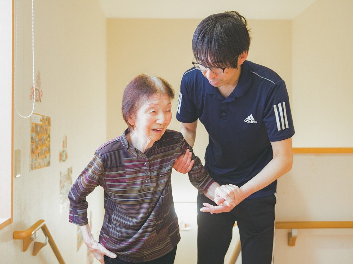 階段の近くで高齢女性の腕を支えて歩いている眼鏡をかけたポロシャツを着た男性スタッフ。